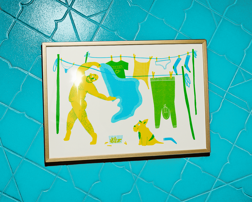 Framed mock up of risograph printed artwork of naked hairy man hanging up washing on line, with a dog unpleasantly looking up at him waving a bed sheet around.